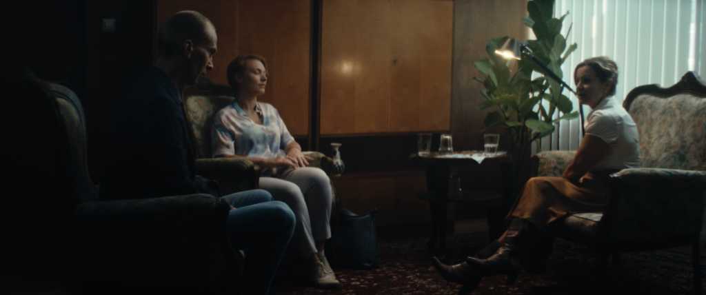 Ein junges Paar und eine Frau mittleren Alters sitzen in einem Raum, der wie ein Therapieraum aussieht. Die ältere Frau sitzt dem Paar gegenüber und sieht die beiden an.