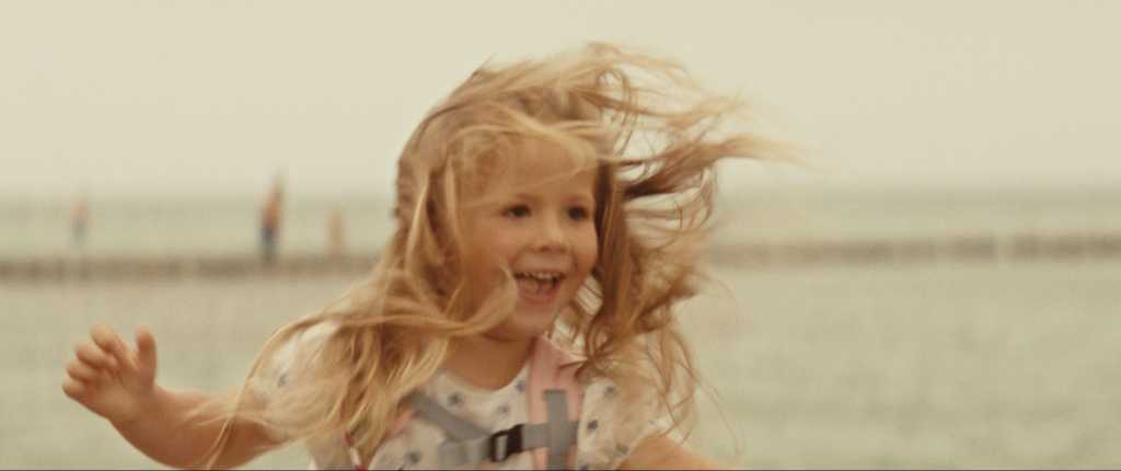 Ein kleines Mädchen steht am Strand in verspielter Pose und lächelt. Ihr Haar ist vom Wind ganz zerzaust.
