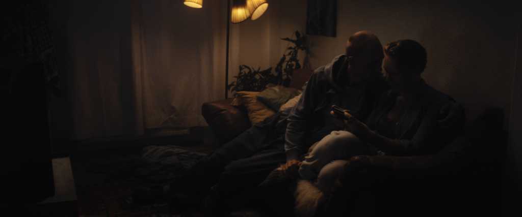 Ein junges Paar sitzt auf einer Couch in einem Raum mit wenig Licht, er versucht, sich ihr zu nähern, sie starrt auf ihr Handy.