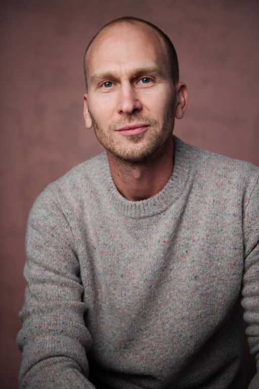 Ein Portraitfoto eines selbstbewusst lächelnden jungen Mannes mit kurzen blonden Haaren, der einen beigen Wollpullover trägt und direkt in die Kamera blickt.